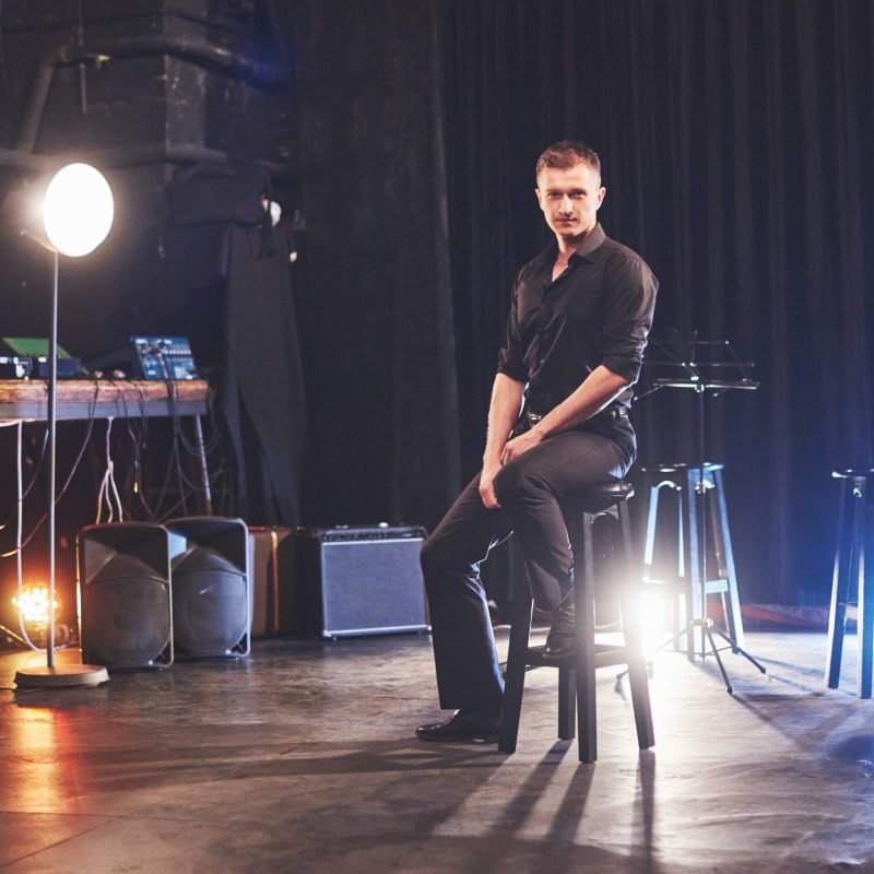 Magic look. Handsome young man in black clothes looking at camera while sitting on chair near the in dark room with light.